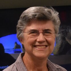 An older white woman with short white hair and frame-less glasses smiles broadly