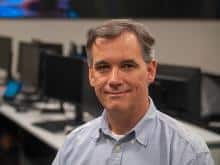 Jerry Hughes; A white man with graying hair smiles at the camera. He wears a light blue button up in front of a row of computers.