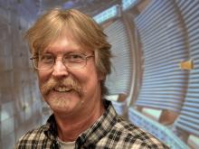 A middle-aged blonde man with a bushy mustache and glasses smiles at the camera